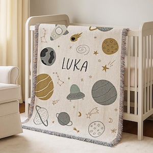 Op de afbeelding: Een crèmekleurige babydeken met een ruimtethema. De deken heeft planeten, sterren, een raket en de naam "LUKA" in zwarte letters. De deken heeft een gefranjerde rand en is over een wit ledikant gedrapeerd.