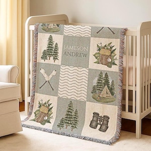 May include: A woven baby blanket with a camping theme. The blanket features squares with images of trees, a tent, binoculars, and crossed campfires. The name "JAMESON ANDREW" is printed on the blanket. The blanket is draped over a white cot.