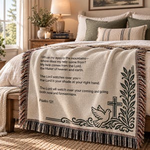 May include: A woven throw blanket in cream with black text featuring Psalm 121. The design includes a dove, a cross, and a leafy branch motif. The blanket has a fringed edge with multi-coloured threads and is draped over a bed with neutral bedding.