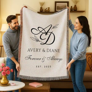 May include: A beige woven throw blanket with a black monogram and text that reads "Avery & Diane Forever & Always EST. 2025". The blanket has a fringed edge and is being held up by two people.