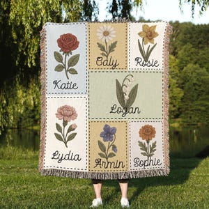 May include: A woven throw blanket with embroidered floral designs in a grid. Each square displays a different flower with a name below, including Katie, Caly, Rosie, Logan, Lydia, Armin, and Sophie. The blanket has a fringed edge.