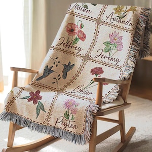 May include: A beige woven throw blanket with a floral and hummingbird design, draped over a wooden rocking chair. The blanket features personalized names and floral illustrations in squares. The blanket has a fringed edge.