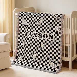 Peut inclure: Une couverture à carreaux noirs et blancs avec le nom "Jaxson" et des éclairs blancs. La couverture est posée sur un lit bébé blanc, avec une bordure à franges.