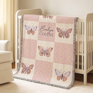 Może przedstawiać: Różowo-kremowy kocyk dla dziecka z motywem motyla i nadrukowanym imieniem "Evelyn Colene". Kocyk jest narzucony na białe łóżeczko. Kocyk ma frędzelki na krawędzi.