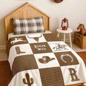 May include: A white and brown patchwork blanket featuring western-themed graphics including a bull skull, cowboy boots, a cowboy on a horse, a hat, a horseshoe, a cactus, and the text RYKER WES, arranged in a grid pattern with polka dot and wavy line squares.