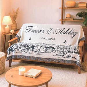 May include: Cream-colored throw blanket with a mountain and tree design, personalized with the names "Trevor & Ashley" and the date "10-07-2023". The blanket has a fringed edge and is draped over a wooden sofa.