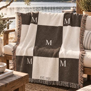 Può includere: Una coperta tessuta con un motivo a scacchi composto da quadrati marrone scuro e crema. Quattro dei quadrati scuri sono ricamati con la lettera M in un carattere serif. Il quadrato centrale inferiore include il testo EST. 2025. Bordi sfrangiati.