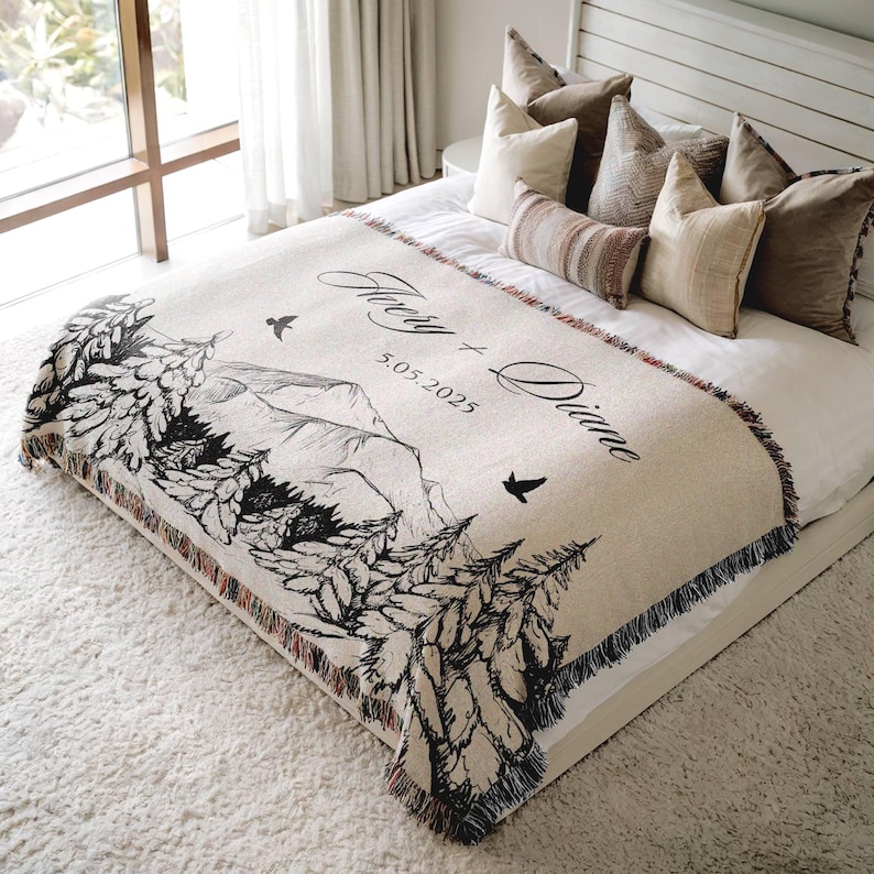 May include: A beige woven blanket with a black and white mountain and tree design. The blanket has the names "Avery + Diane" and the date "5.05.2025" printed on it. The blanket is fringed on the edges and is displayed on a bed.