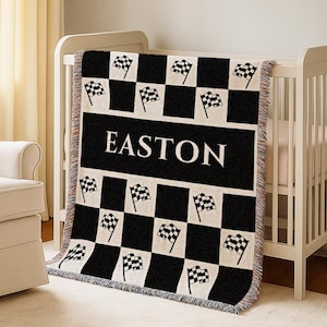 Op de afbeelding: Een zwart-wit geblokte babydeken met de naam "EASTON" in witte blokletters. De deken heeft racevlaggen en is over een witte wieg gedrapeerd. De deken heeft een gefranjerde rand.