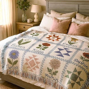 May include: Cream-coloured throw blanket with a patchwork design featuring floral and geometric patterns. Each square has a name and a flower illustration. The blanket has a fringed edge and is on a bed with pillows.