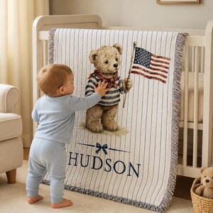 May include: A woven throw blanket featuring a light cream background with thin vertical blue stripes. The center displays a brown teddy bear wearing a blue and white striped shirt and a red scarf, holding an American flag. The name HUDSON is printed below.