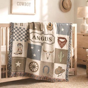 Könnte beinhalten: Eine gewebte Decke mit Cowboy-Motiv, mit Quadraten mit einer Gitarre, einem Cowboy-Stiefel, einem Stierschädel und einem Hufeisen. Die Decke ist über ein Kinderbett drapiert und trägt in der Mitte den Namen "ANGUS". Die Decke hat einen Fransenrand.