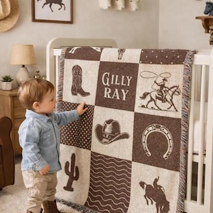 May include: A woven throw blanket featuring a brown and cream checkerboard pattern with western-themed graphics including a cowboy boot, a cowboy hat, a horseshoe, a cactus, and a rider with a lasso, with the name Gilly Ray printed in the center.