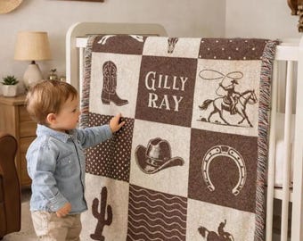 May include: A woven throw blanket featuring a brown and cream checkerboard pattern with western-themed graphics including a cowboy boot, a cowboy hat, a horseshoe, a cactus, and a rider with a lasso, with the name Gilly Ray printed in the centre.
