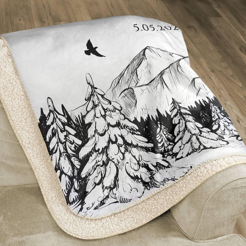 May include: A white and cream-colored blanket with a black and white mountain and forest design. The blanket has a soft, plush texture and features a drawing of a bird and the date 5.05.202. The blanket is folded over a beige sofa.