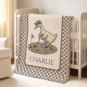Könnte beinhalten: Eine beige-braune Babydecke mit einer Cartoon-Gans, die Golf spielt. Die Decke hat einen Vichy-Karo-Rand und den Namen "CHARLIE" aufgedruckt. Die Decke ist über ein weißes Babybett gelegt.