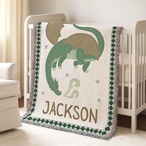 Könnte beinhalten: Eine cremefarbene Babydecke mit einem grünen Drachendesign und dem Namen "JACKSON" in braunen Buchstaben. Die Decke hat einen grün-braunen Rand mit Fransen. Sie liegt über einem weißen Kinderbett.