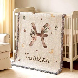 May include: A beige baby blanket with a Pisces symbol and the name "Dawson" embroidered in a script font. The blanket features floral and celestial accents, with a fringed edge. The blanket is draped over a white crib.