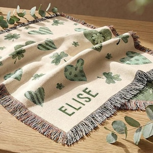 May include: A beige throw blanket with a green heart and clover pattern. The word "ELISE" is printed in green on the bottom corner. The blanket has a fringed edge with a mix of colors.