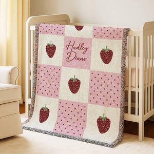 May include: A woven baby blanket with a strawberry theme. The blanket features a patchwork design with alternating squares of strawberries, polka dots, and the name "Hadley Diane" in a script font. The blanket is pink, white, and red.
