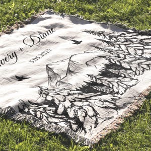 May include: A cream-colored woven throw blanket with a black mountain and tree design. The blanket has a fringed edge and features the names "Avery + Diane" and the date "5.05.2025" in black script.