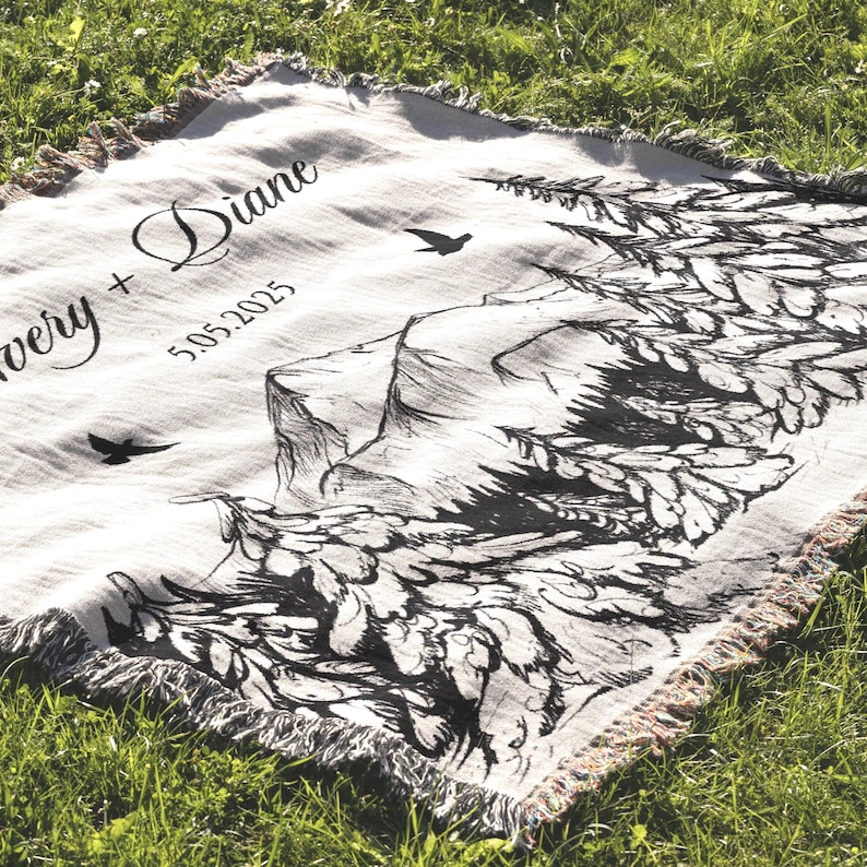 May include: A cream-colored woven throw blanket with a black mountain and tree design. The blanket has a fringed edge and features the names "Avery + Diane" and the date "5.05.2025" in black script.