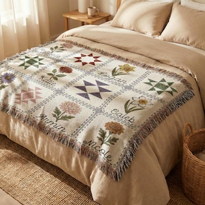 May include: A beige throw blanket with a patchwork design featuring floral and geometric patterns. The blanket has a fringed edge and includes names like "Katie" and "Logan". The blanket is displayed on a bed with beige bedding.