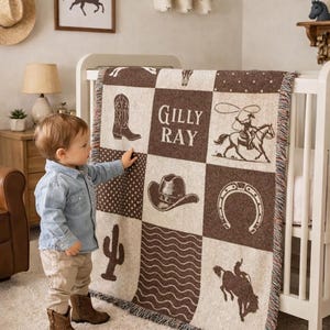 May include: A woven throw blanket featuring a brown and cream checkerboard pattern with western-themed illustrations including a cowboy boot, a cowboy hat, a horseshoe, a cactus, a lassoing cowboy, a bucking horse, and the name Gilly Ray.