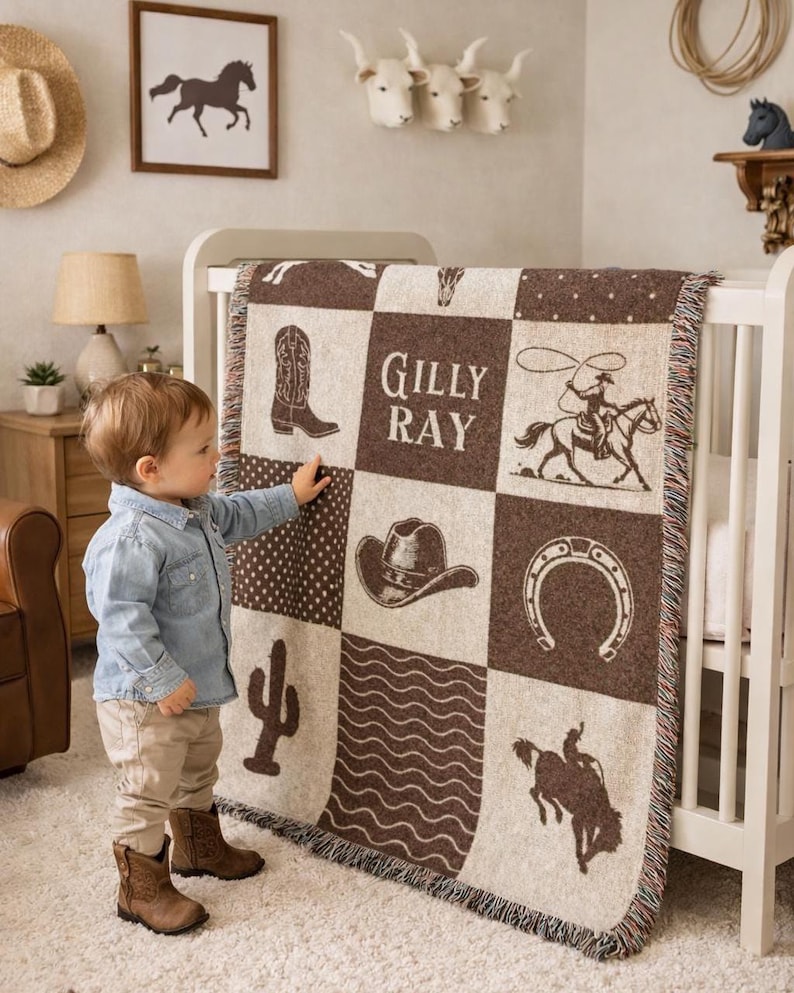 May include: A woven throw blanket featuring a brown and cream checkerboard pattern with western-themed illustrations including a cowboy boot, a cowboy hat, a horseshoe, a cactus, a lassoing cowboy, a bucking horse, and the name Gilly Ray.