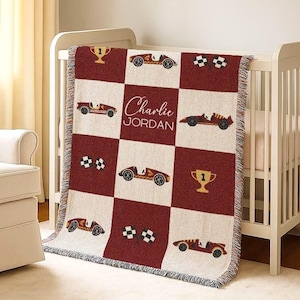 May include: A woven baby blanket with a cream and burgundy checkered design. The blanket features race cars, checkered flags, and a trophy design. The name "Charlie Jordan" is displayed in the centre.