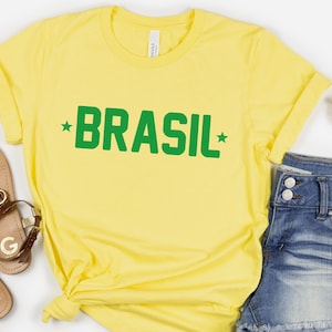 Puede incluir: Camiseta amarilla con texto verde que dice "BRASIL" con una estrella a cada lado de la palabra.