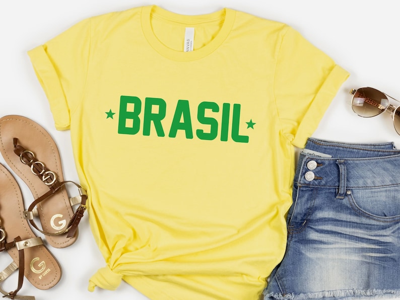 Puede incluir: Camiseta amarilla con texto verde que dice "BRASIL" con una estrella a cada lado de la palabra.