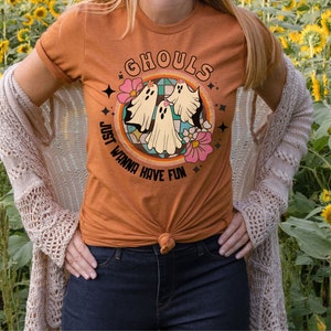 May include: A woman wearing a burnt orange t-shirt with a retro-style graphic of three ghosts and the text "Ghouls Just Wanna Have Fun". She is standing in a field of sunflowers.