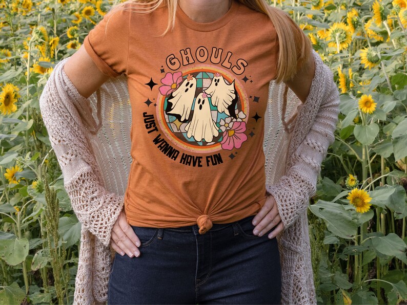 May include: A woman wearing a burnt orange t-shirt with a retro-style graphic of three ghosts and the text "Ghouls Just Wanna Have Fun". She is standing in a field of sunflowers.