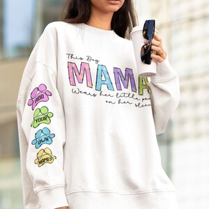 K&ouml;nnte beinhalten: Wei&szlig;er Pullover mit dem Text "This Dog Mama Wears Her Little Paws On Her Sleeve" und einem bunten Pfotenabdruck-Design auf dem &Auml;rmel. Der Text "Add Your Own Names" ist auf dem &Auml;rmel gedruckt.