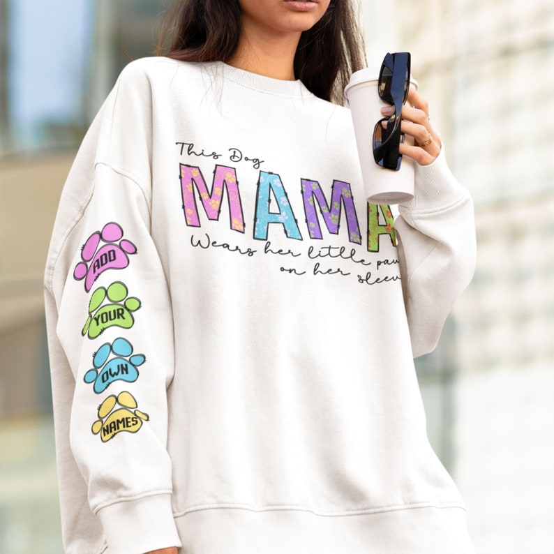 K&ouml;nnte beinhalten: Wei&szlig;er Pullover mit dem Text "This Dog Mama Wears Her Little Paws On Her Sleeve" und einem bunten Pfotenabdruck-Design auf dem &Auml;rmel. Der Text "Add Your Own Names" ist auf dem &Auml;rmel gedruckt.