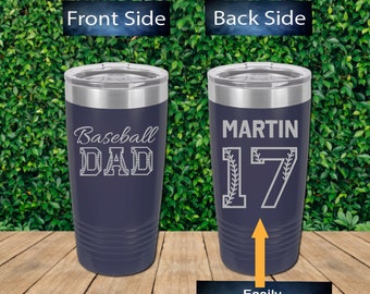 May include: A navy blue stainless steel tumbler with a silver lid. The front of the tumbler is engraved with the text "Baseball Dad". The back of the tumbler is engraved with the text "MARTIN 17" with a baseball stitch design around the number.