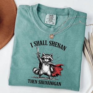 Puede incluir: Una camiseta Comfort Colors de color turquesa con una ilustración de un mapache con una capa roja y una espada. El texto dice "I SHALL SHENAN THEN SHENANIGAN". Un collar plateado con un colgante está sobre la camiseta.