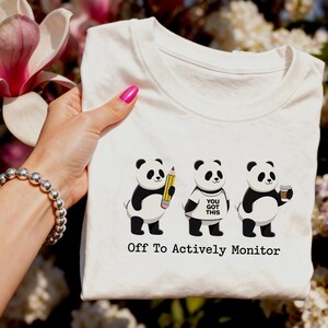 Peut inclure: T-shirt blanc avec trois pandas dessinés. L'un tient un crayon, un autre porte un t-shirt avec l'inscription "You Got This", et le troisième tient une tasse de café. Le texte "Off To Actively Monitor" est en dessous.
