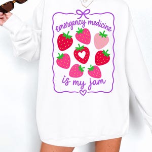 Peut inclure: Un sweat-shirt blanc à manches longues avec un cadre rectangulaire violet orné d'un nœud en haut et d'un cœur en bas. À l'intérieur, des fraises rouges et roses accompagnent le texte violet "emergency medicine is my jam".