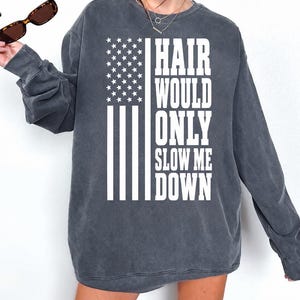 Peut inclure: Un sweat-shirt à col rond gris anthracite avec une impression graphique blanche représentant un drapeau américain et le texte "HAIR WOULD ONLY SLOW ME DOWN" en lettres capitales grasses. Le vêtement a des manches longues et une coupe ample.