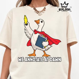 May include: A cream-colored t-shirt featuring a cartoon goose wearing a red cape, holding a yellow highlighter and a blue book. The text "WE ANNOTATE AT DAWN" is printed below the goose, and the "EXLOOM" logo is visible in the top right corner.