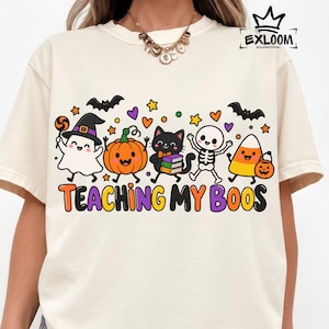 Peut inclure: Un t-shirt couleur crème avec un motif d'Halloween coloré représentant un fantôme, une citrouille, un chat noir, un squelette et un bonbon maïs. Le texte "TEACHING MY BOOS" est écrit en lettres grasses multicolores avec des chauves-souris.