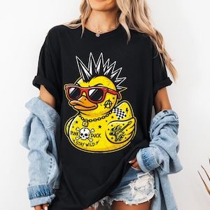 Peut inclure: T-shirt noir avec un motif de canard punk jaune. Le canard porte des lunettes de soleil rouges, une chaîne et une crête iroquoise. Le graphisme comprend une tête de mort, une guitare et le texte "Punk Duck Stay Wild". Le design est vieilli.