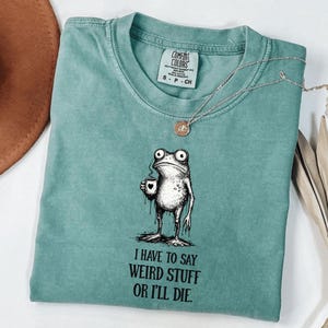 Op de afbeelding: Een turquoise Comfort Colors T-shirt met een illustratie van een kikker die een koffiekopje vasthoudt. De kikker heeft een vreemde uitdrukking. De tekst "I HAVE TO SAY WEIRD STUFF OR I'LL DIE" staat eronder. Een zilveren ketting ligt op het shirt.