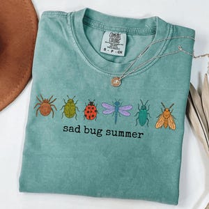 May include: A sage green short-sleeve t-shirt featuring a graphic print of six colourful insects including a spider, beetle, ladybug, dragonfly, and moth. Below the insects, the text reads "sad bug summer" in black, lowercase, sans-serif font.