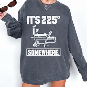 Peut inclure: Un sweat-shirt gris anthracite à col rond avec une impression graphique blanche représentant un fumoir à barbecue avec de la fumée s'échappant de la cheminée. Le texte au-dessus indique IT'S 225° et le texte en dessous SOMEWHERE dans un cadre.