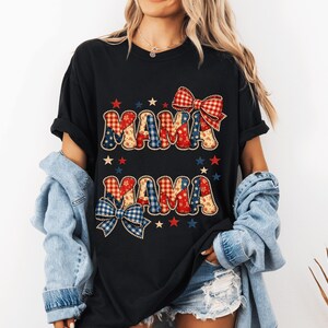 Peut inclure: T-shirt noir avec le mot "MAMA" dans un motif patriotique. Les lettres sont remplies de motifs rouges, blancs et bleus, et le motif comprend des étoiles et des nœuds. Un t-shirt décontracté et confortable pour un usage quotidien.