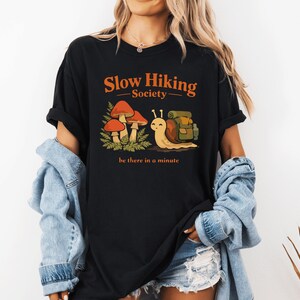 Peut inclure: T-shirt noir avec l'inscription orange "Slow Hiking Society". Le t-shirt présente un escargot de dessin animé avec un sac à dos, des champignons et la phrase "be there in a minute". Le t-shirt est de style décontracté et confortable.