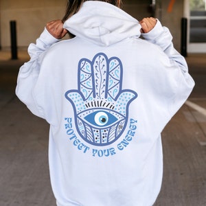 May include: White hoodie with a large blue Hamsa hand design on the back, featuring an eye symbol and the text "PROTECT YOUR ENERGY" in a matching blue color. The hoodie has a drawstring hood and long sleeves.